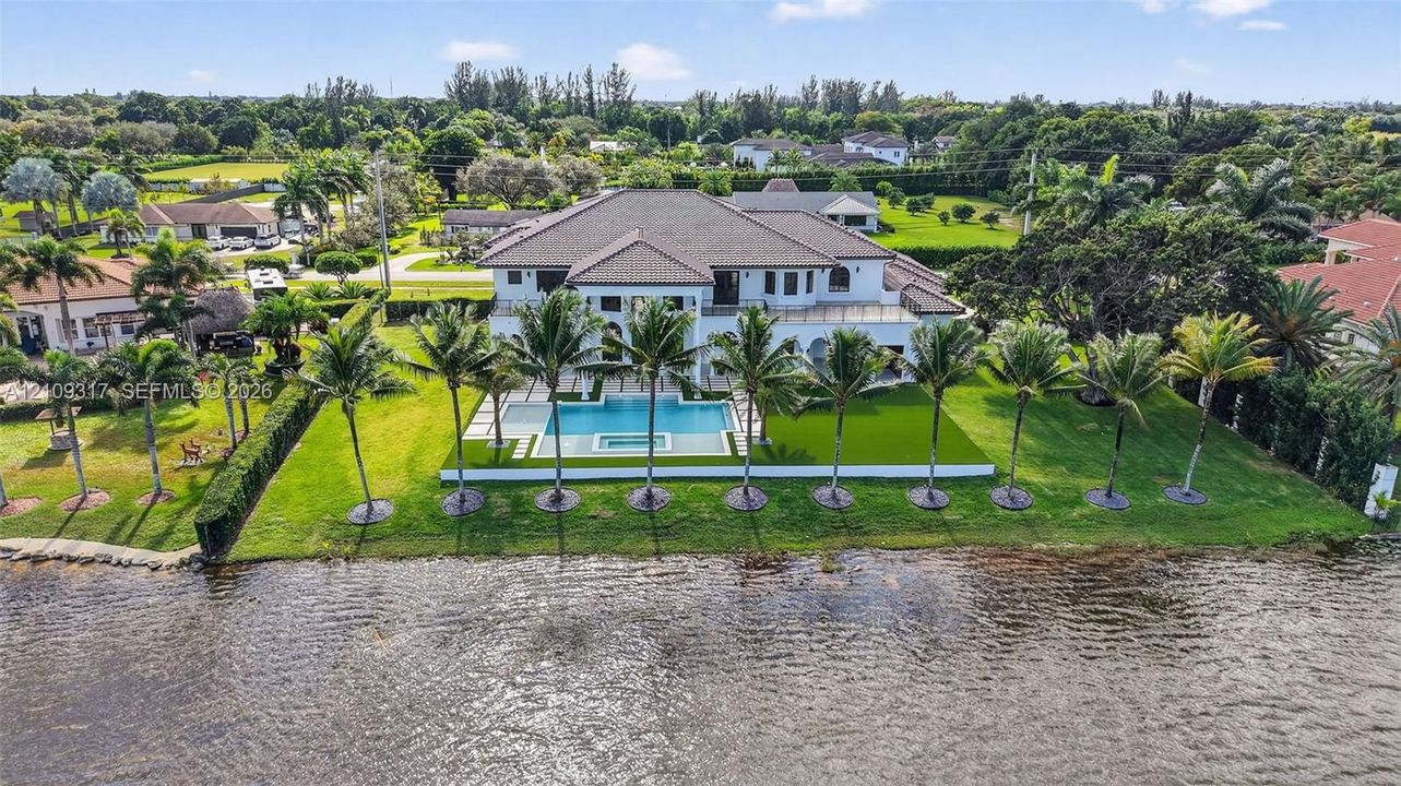 Waterfront estate featuring a multi-level roofline, swimming pool with integrated spa, paved terrace, and extensive manicured landscaping with mature palm trees