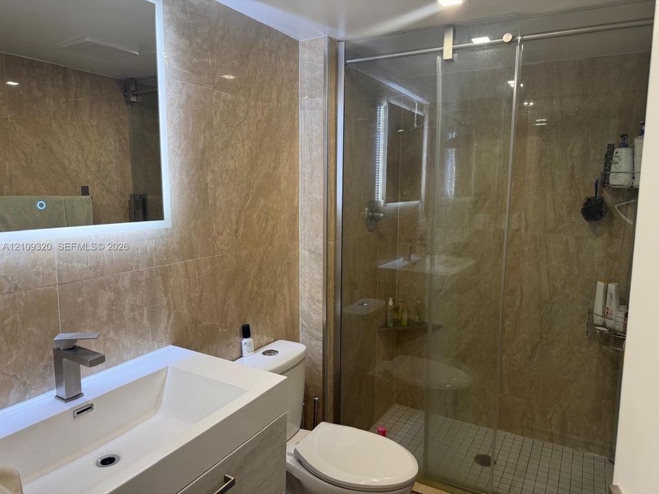 Full-height wall tiling featuring natural stone-patterned surfaces, a wall-mounted LED-backlit mirror, a white rectangular vessel sink with a single-handle chrome faucet, and a glass-enclosed shower with tiled flooring