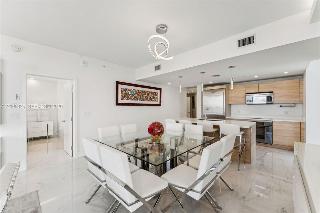Open-concept kitchen and dining area featuring light-toned wood-finish cabinetry, white stone-composite countertops, stainless steel appliances, large-format polished tile flooring, and contemporary overhead lighting fixtures