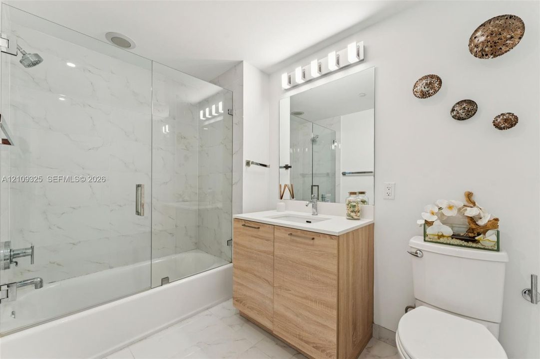 Bathroom featuring a glass-enclosed shower and tub combination with wall-to-wall surfacing, a vanity with wood-finish cabinetry, a white countertop with an integrated sink, and an expansive vanity mirror with overhead lighting