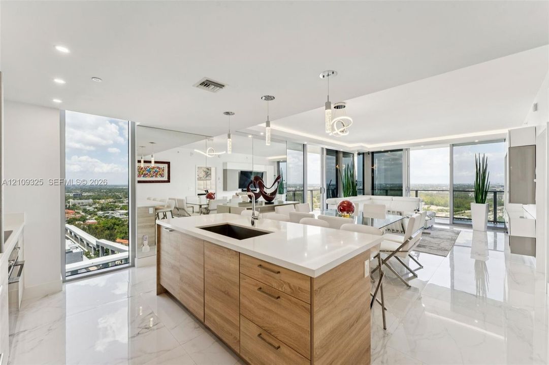 Kitchen island with wood-finish cabinetry, white countertop, and under-mount sink, complemented by polished tile flooring, floor-to-ceiling glass panels, and recessed and pendant lighting