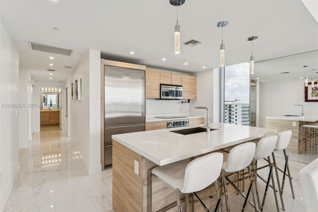 Modern kitchen featuring light wood-finish cabinetry, a central island with a quartz countertop, stainless steel refrigerator, recessed lighting, and polished stone-finish flooring