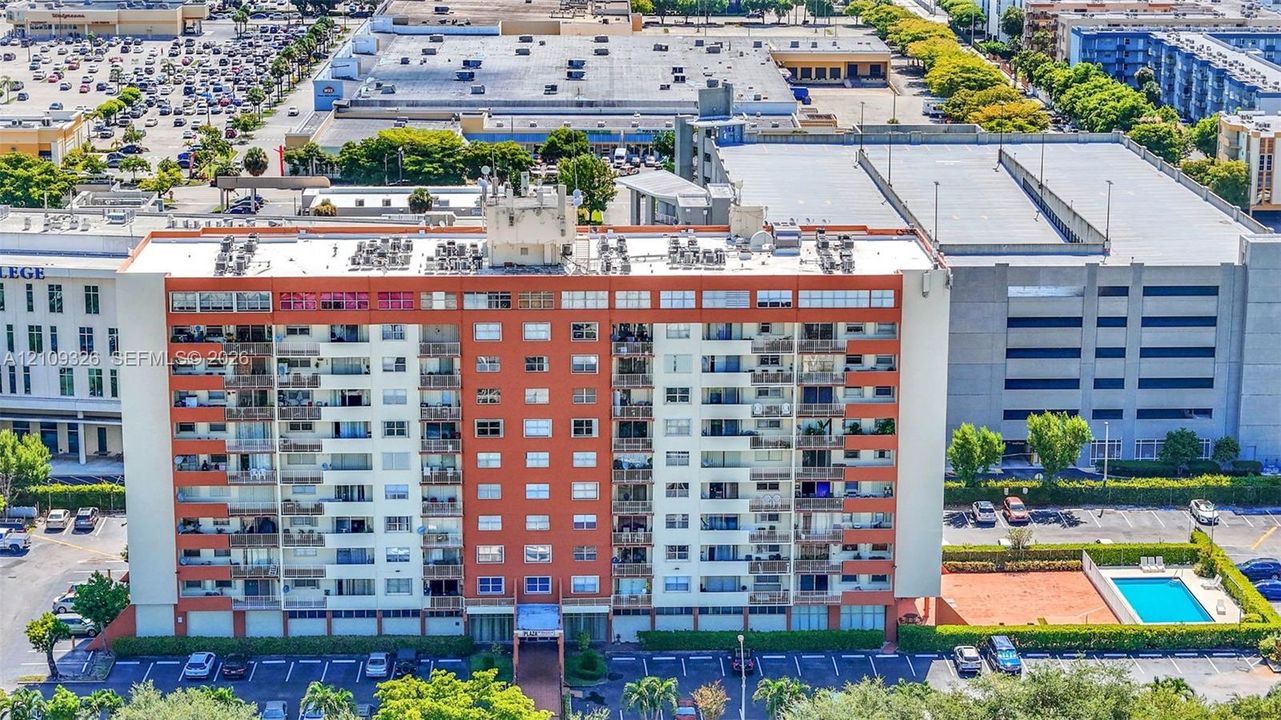 Mid-rise residential building featuring multiple private balconies, a ground-level swimming pool, and an asphalt parking lot