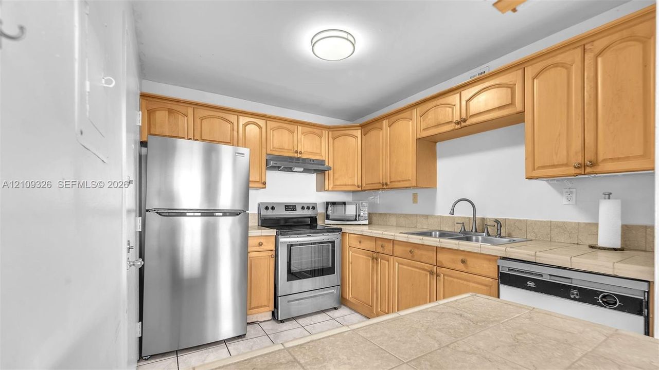 Kitchen featuring natural wood-finish cabinetry, tile countertops, stainless steel appliances, and tile flooring