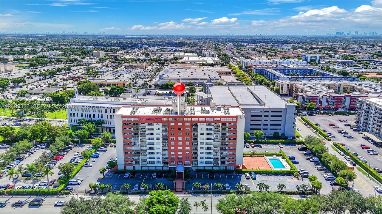 Aerial perspective of a multi-story residential building featuring private balconies, an on-site swimming pool, paved surface parking, and manicured landscaping