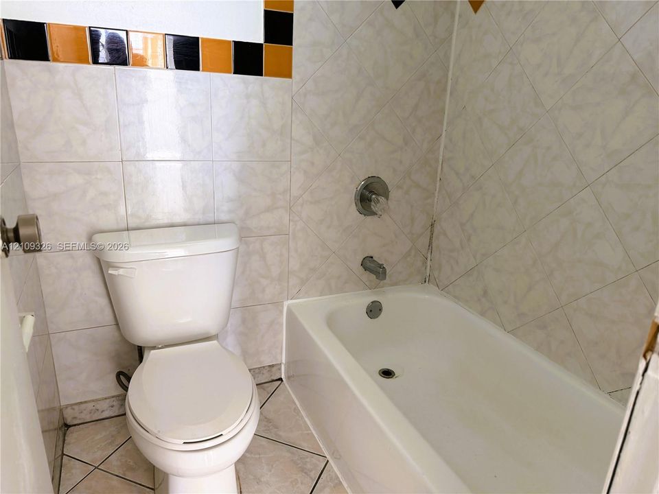Bathroom featuring a white porcelain commode, a composite bathtub with chrome plumbing fixtures, and wall tiling accented by a decorative border