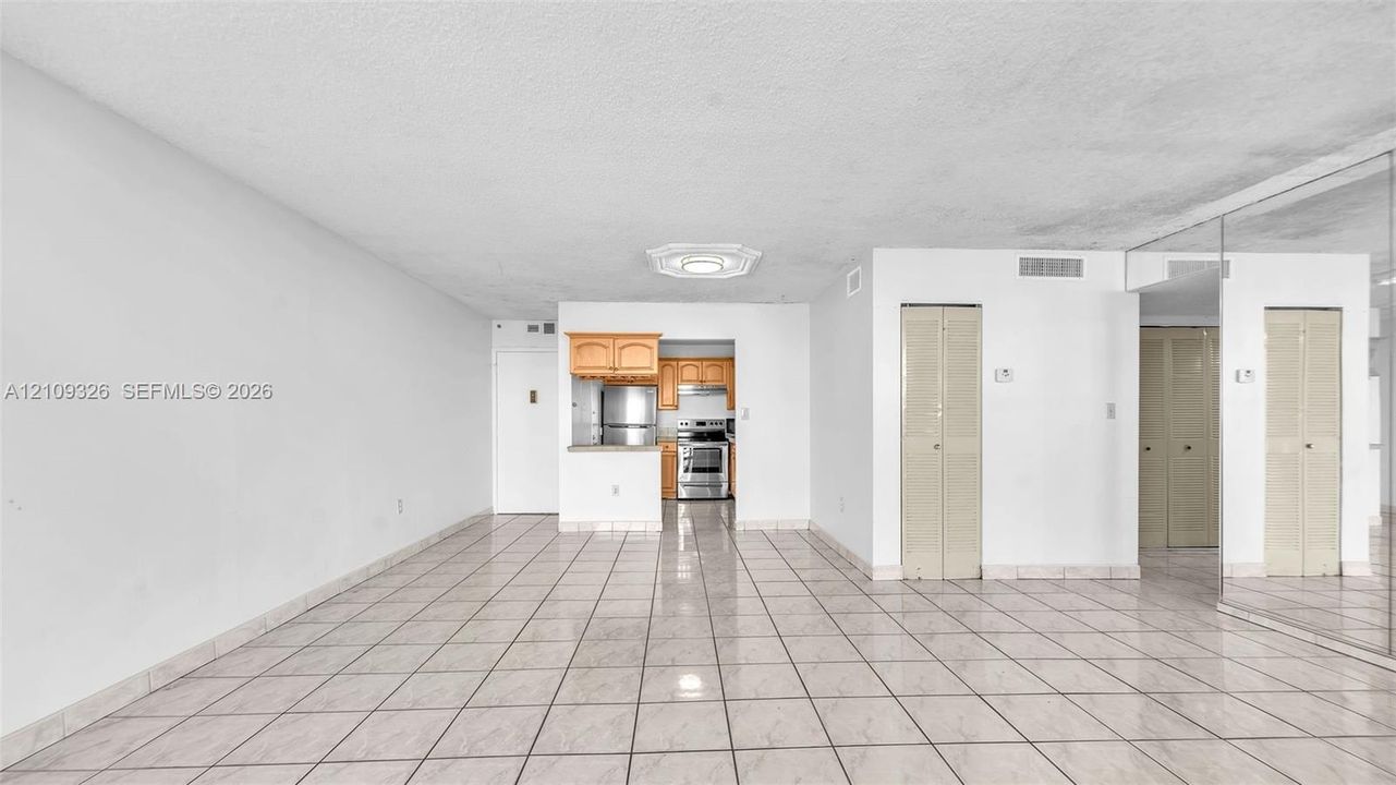 Open-concept interior featuring neutral tile flooring, mirrored wall paneling, built-in bi-fold closet doors, and recessed ceiling lighting, with access to a kitchen area equipped with stainless steel appliances and wood cabinetry