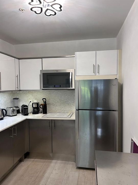 Kitchen featuring two-tone cabinetry with white upper and stainless steel lower doors, stainless steel refrigerator and built-in microwave, solid surface countertops, mosaic tile backsplash, and light-toned wood-finish flooring