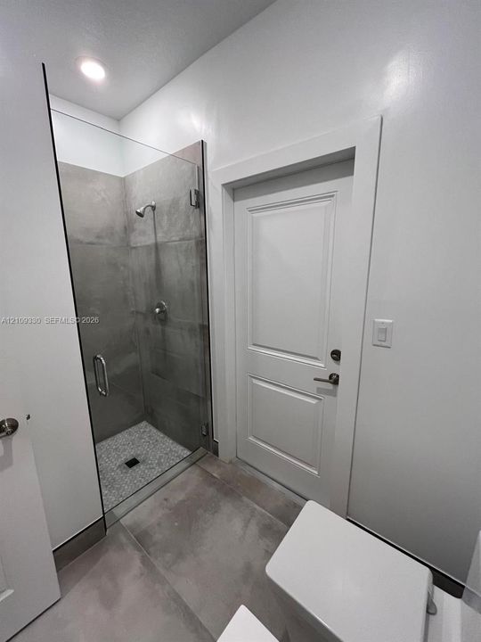 Walk-in shower featuring a frameless glass door, large-format grey tile surround, mosaic stone-finish floor, and metallic hardware