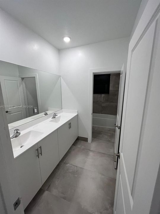 Dual-sink vanity with white cabinetry, sleek hardware, stone countertop, large wall-to-wall mirror, and oversized tile flooring