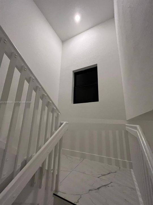 Stairwell featuring white vertical balusters, light-colored tile flooring with distinctive veining, recessed ceiling lighting, and a square window