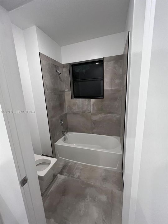 Full-height tiled walls surrounding a soaking tub with integrated shower hardware, large-format tile flooring, and a window with a black frame