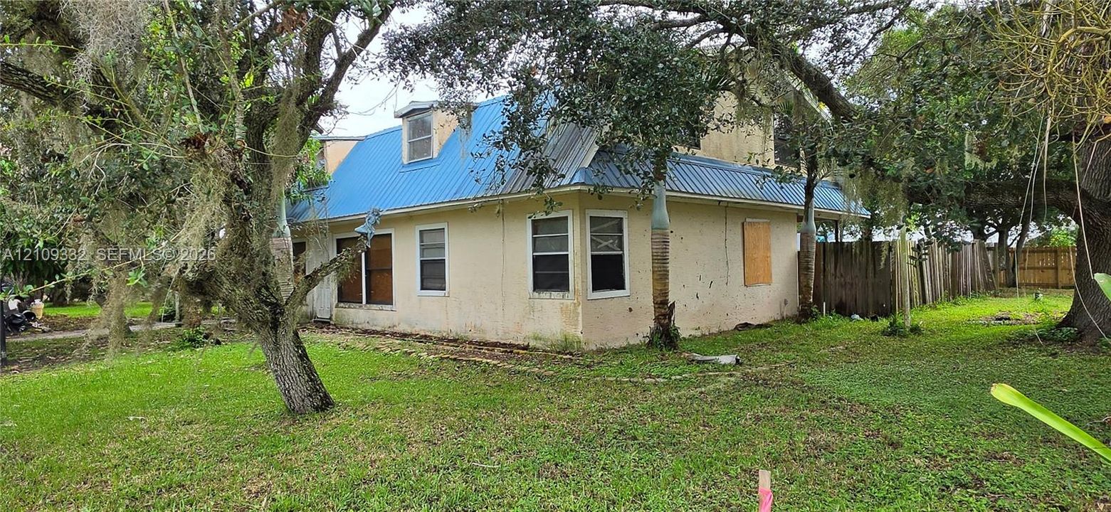 Exterior featuring a light-colored stucco facade, blue standing-seam metal roofing, dormer window, and mature trees on the surrounding grounds