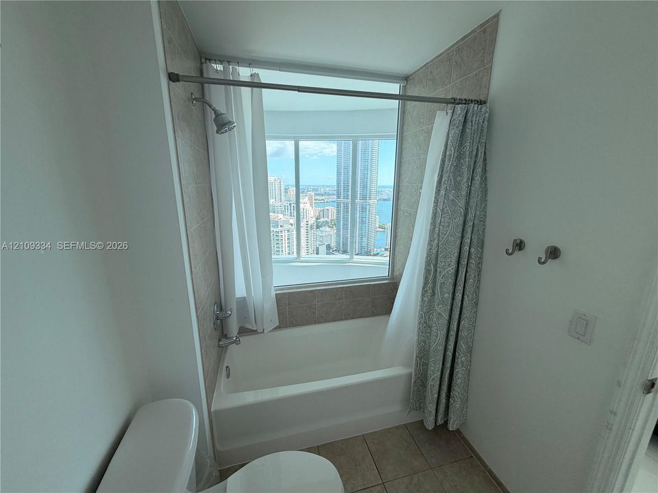 Bathroom featuring a built-in bathtub, light-toned ceramic tile surround, integrated shower fixture, and a large window with an expansive urban skyline and water backdrop