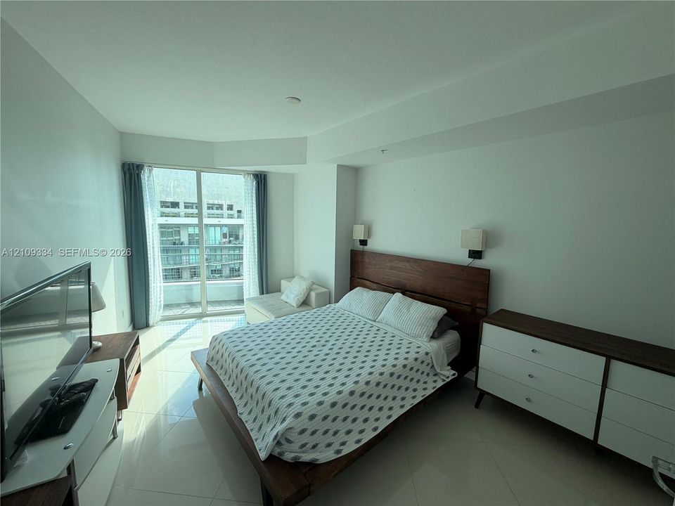 Floor-to-ceiling glass door, polished tile flooring, wall-mounted bedside light fixtures, and recessed architectural ceiling detailing
