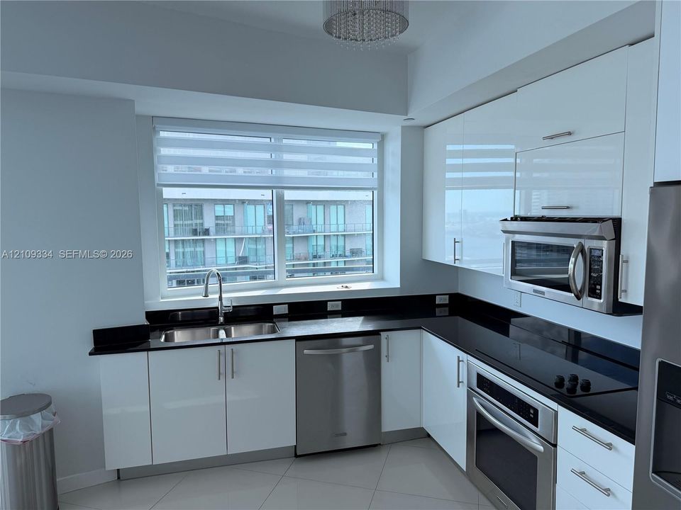 Kitchen featuring high-gloss white cabinetry, dark stone-finish countertops, stainless steel appliances, large double-basin under-mount sink, and a modern drum-style ceiling fixture