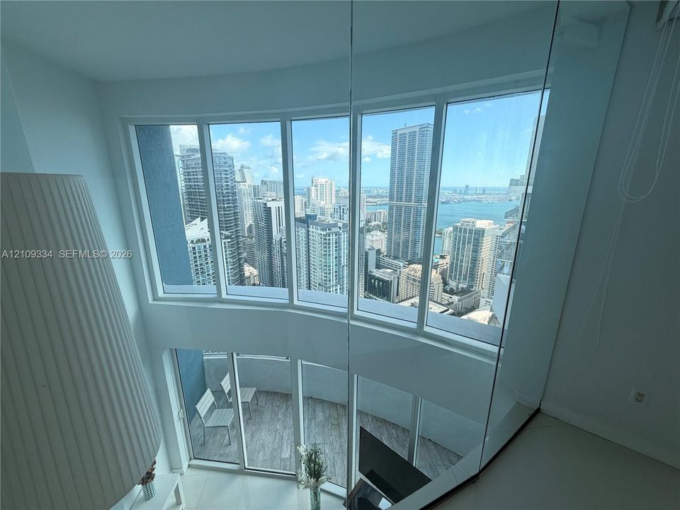 Double-height floor-to-ceiling windows featuring city and water scenery, glass partition railing, and private balcony with wood-finish flooring