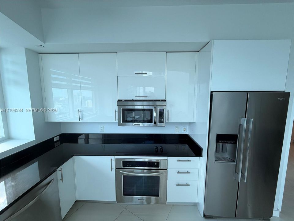 Contemporary kitchen featuring high-gloss white cabinetry, dark stone countertops, stainless steel appliances, and large-format tile flooring