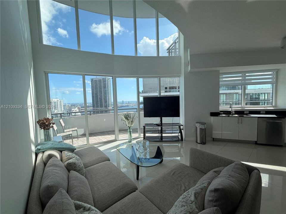 Double-height floor-to-ceiling windows, polished tile flooring, glass balcony enclosure, and wet bar featuring white cabinetry with a dark countertop