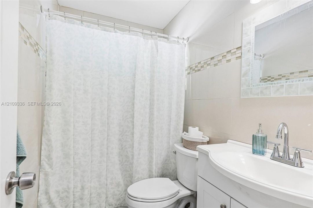 Bathroom featuring full-height wall tiling with mosaic accent banding, white vanity cabinet with an integrated sink, polished chrome faucet, and a wall-mounted mirror with a tiled frame