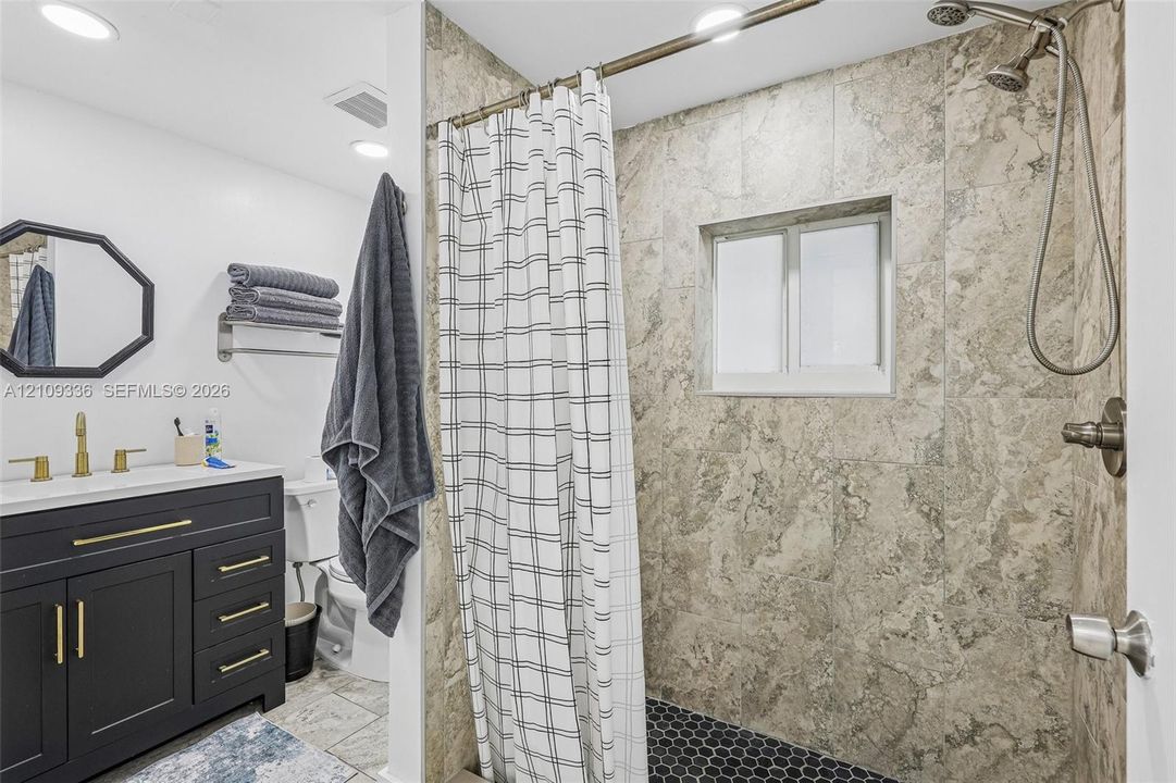 Bathroom featuring a dark-finish vanity with gold-tone hardware, stone-surround shower with a handheld fixture, hexagonal tile shower floor, and a framed wall mirror