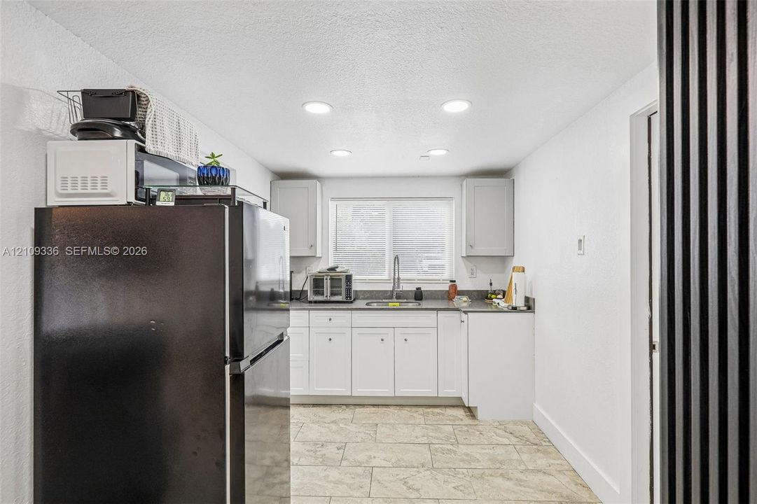 Kitchen featuring white shaker-style cabinetry, solid-surface countertops, stone-finish tile flooring, recessed ceiling lighting, and a stainless steel kitchen sink
