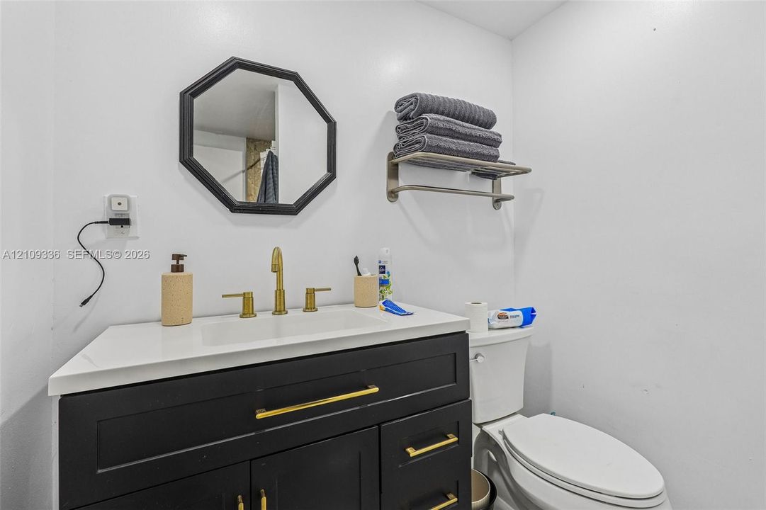 Dark cabinetry vanity with gold-tone hardware, rectangular white countertop basin, brushed gold widespread faucet, wall-mounted octagonal mirror, and metal wall-mounted shelving