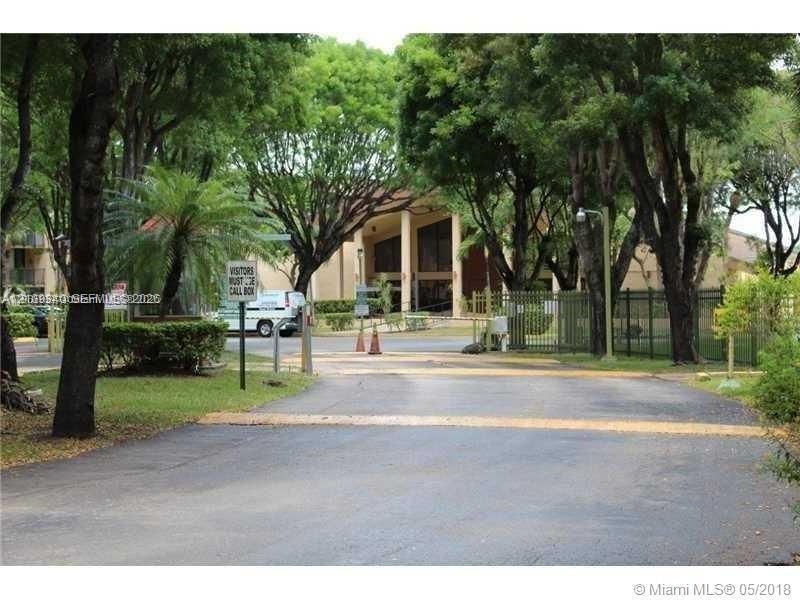 Paved entrance featuring mature canopy trees, metal perimeter fencing, and an gated access point