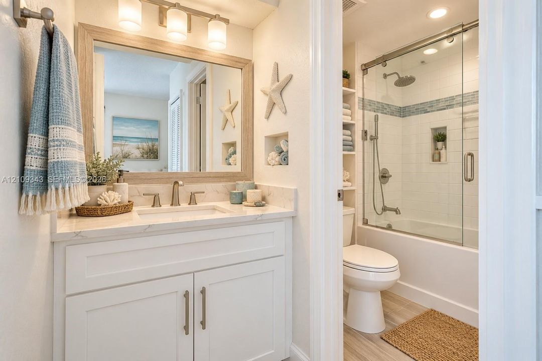 Bathroom featuring a white shaker-style vanity with stone-finish countertop, undermount sink, framed mirror with three-light fixture, built-in shelving, and a tub-shower combination with sliding glass doors and recessed wall niche