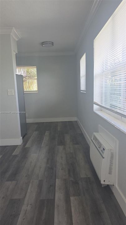 Wood-finish flooring, neutral wall tones, crown molding, windows with integrated blinds, and wall-mounted climate control unit