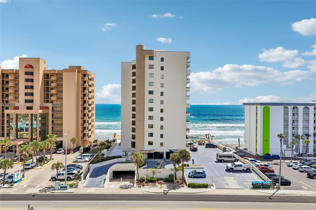 High-rise beachfront building featuring a multi-story exterior, paved parking area, ground-level covered parking access, and mature palm trees