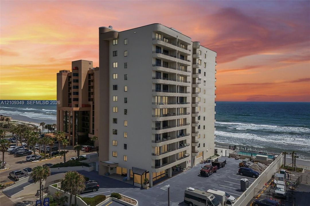 High-rise residential building featuring multiple private balconies, a concrete parking lot, and proximity to the coastline