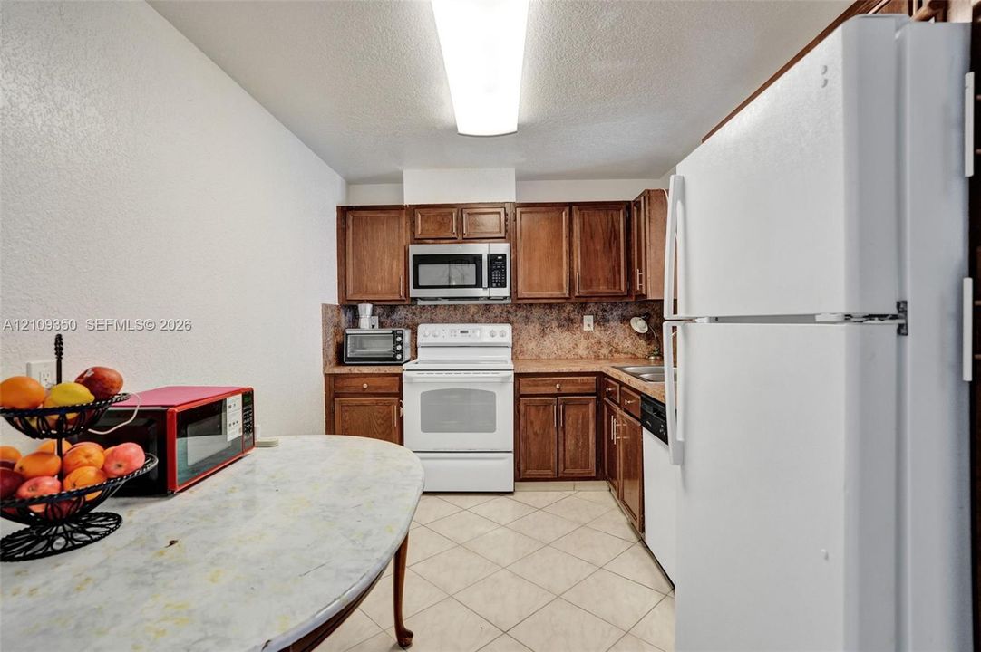 Kitchen featuring wood cabinetry, solid-surface countertops, tile flooring, and integrated white appliances including a range, microwave, and dishwasher
