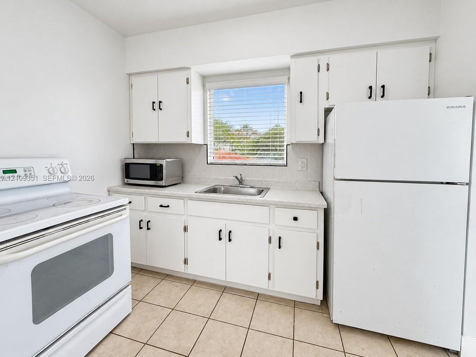 Kitchen featuring white cabinetry with black hardware, stone-composite countertops, stainless steel sink, tile flooring, and integrated white appliances