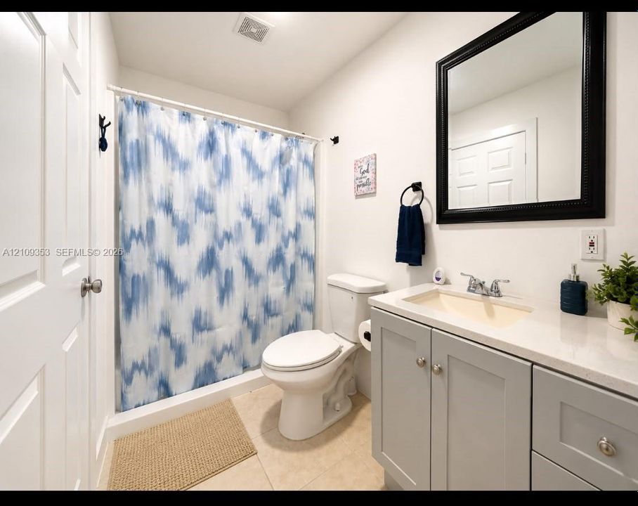 Bathroom featuring a vanity with light gray shaker-style cabinetry, a solid surface countertop, an integrated rectangular sink, a black-framed mirror, and a neutral-toned tile floor