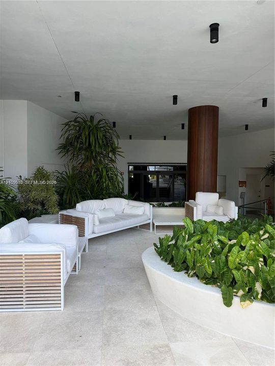 Covered outdoor lounge area featuring large-format stone tile flooring, recessed ceiling lighting, a prominent architectural column with wood-finish cladding, and a curved concrete planter