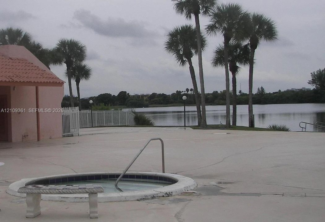 Circular inground spa with a metal handrail, concrete deck, and perimeter white fencing situated on a waterfront lot with mature palm trees