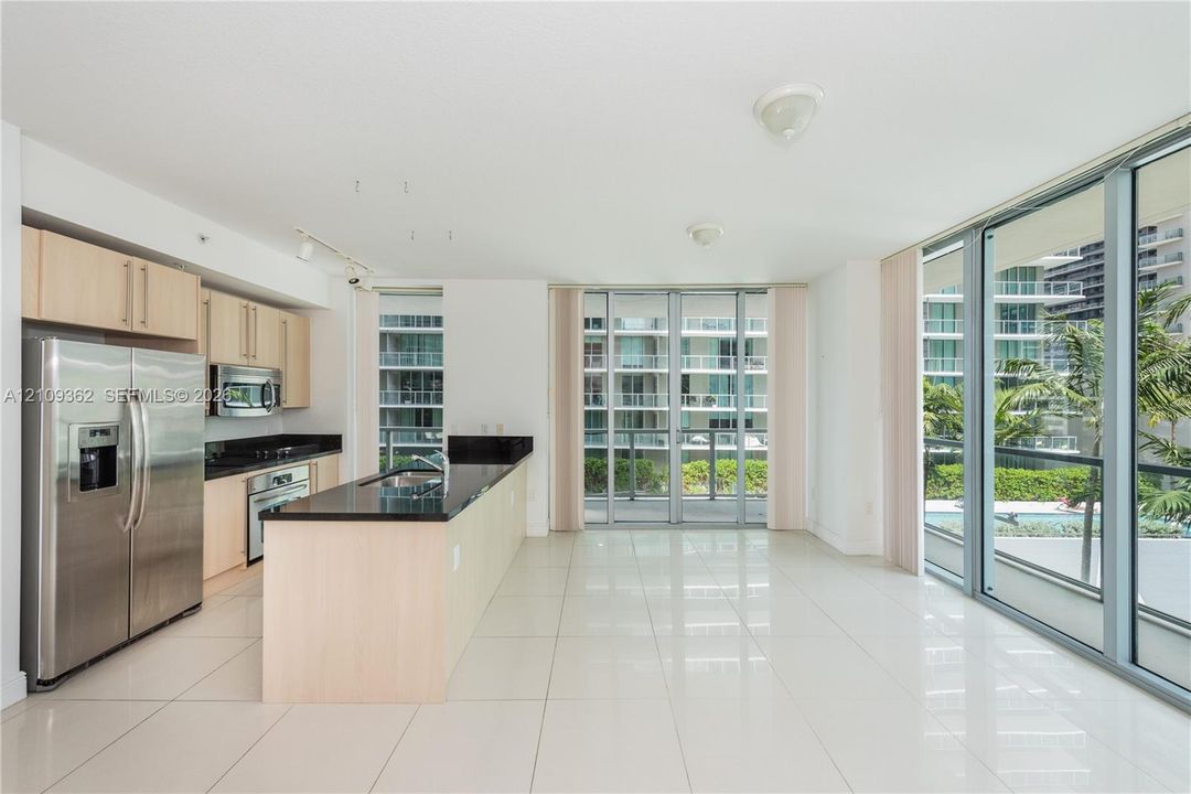 Kitchen area featuring light-finish cabinetry, dark solid-surface countertops, stainless steel appliances, floor-to-ceiling glass sliding doors, and light-toned tile flooring