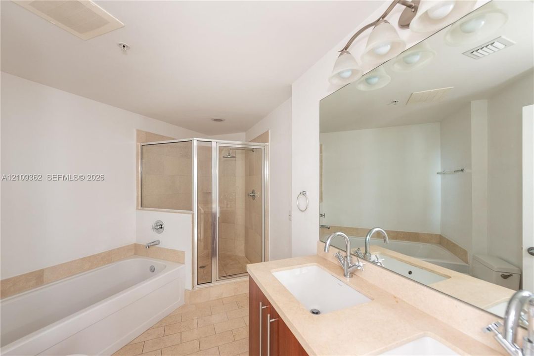 Bathroom featuring a wood-finish double vanity with a light-tone countertop, an expansive wall-to-wall mirror, a soaking tub with tile surround, and a glass-enclosed shower with tile wall finish
