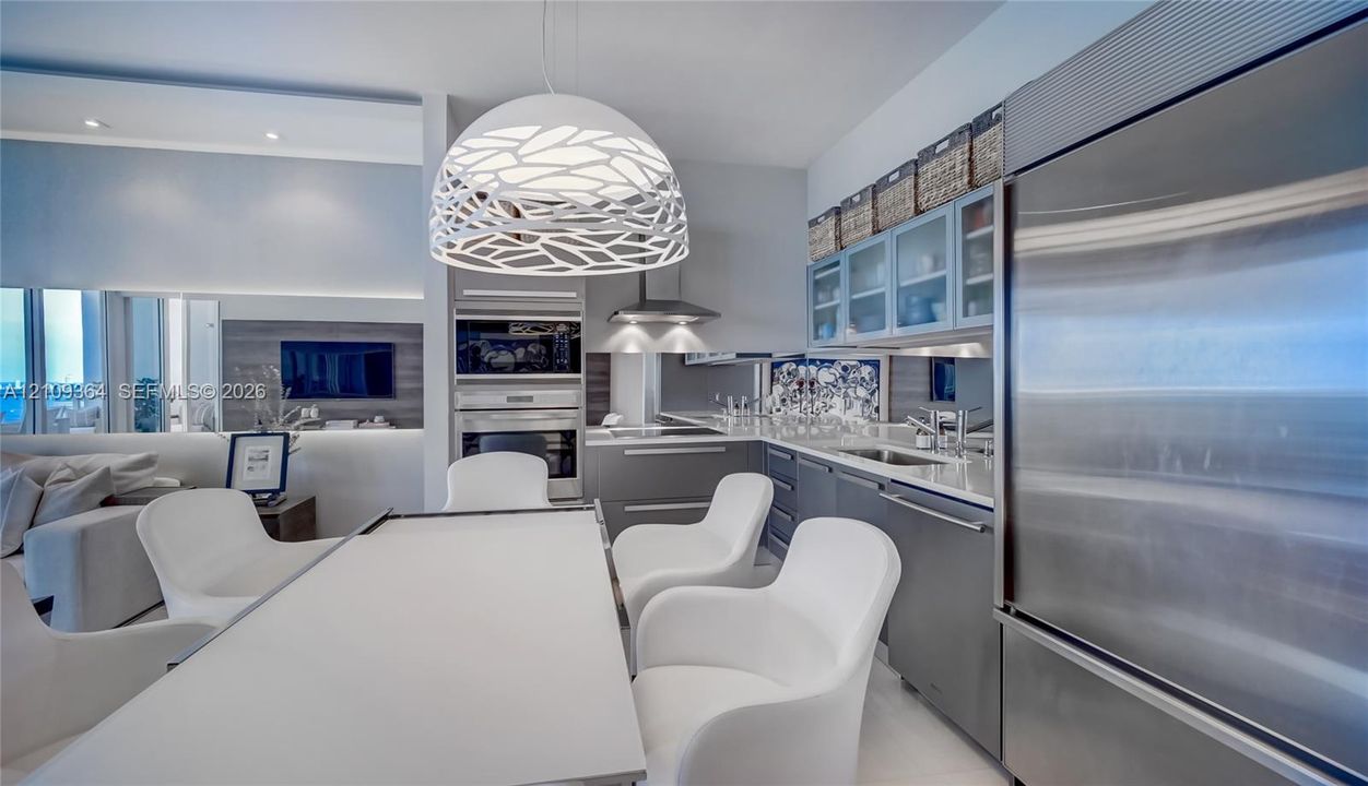 Kitchen featuring stainless steel appliances, built-in dual wall ovens, custom cabinetry with glass-front uppers, stone countertops, and a modern statement pendant fixture