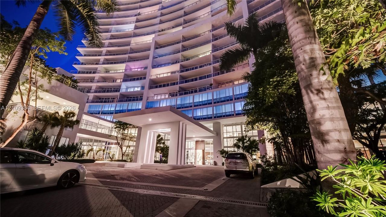 High-rise building exterior featuring a curved facade, multi-story glass-railed balconies, a grand covered porte-cochère entry, and paved motor court