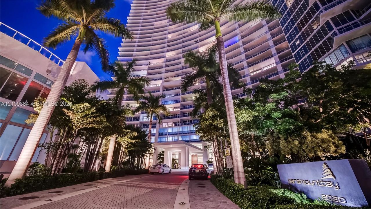 High-rise architectural exterior featuring tiered glass balconies, paved motor court, mature palm landscaping, and structural entryway