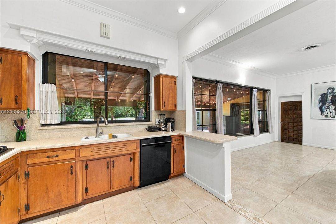 Kitchen featuring warm-tone wood cabinetry, tile flooring, a dual-basin sink with a gooseneck faucet, a built-in black dishwasher, and expansive windows providing natural light
