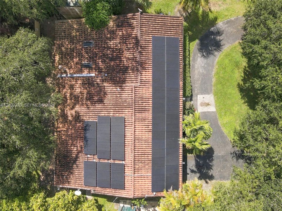 Aerial view of a tile roof featuring integrated solar panel arrays and roof-mounted skylights, set upon a lot with a paved driveway and mature landscaping