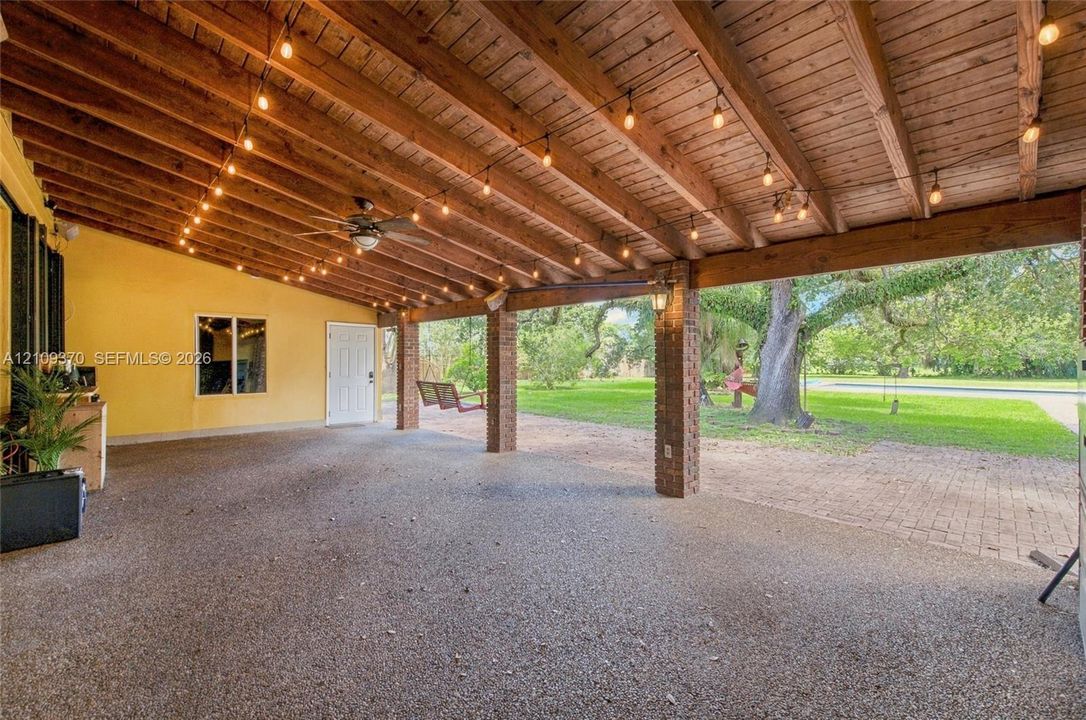 Covered patio featuring an exposed wood beam ceiling, brick support columns, aggregate concrete flooring, and string lighting