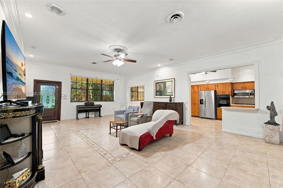 Open-concept living space featuring large-format tile flooring, crown molding, a central ceiling fan with integrated lighting, recessed lighting, and a kitchen pass-through window with a breakfast bar