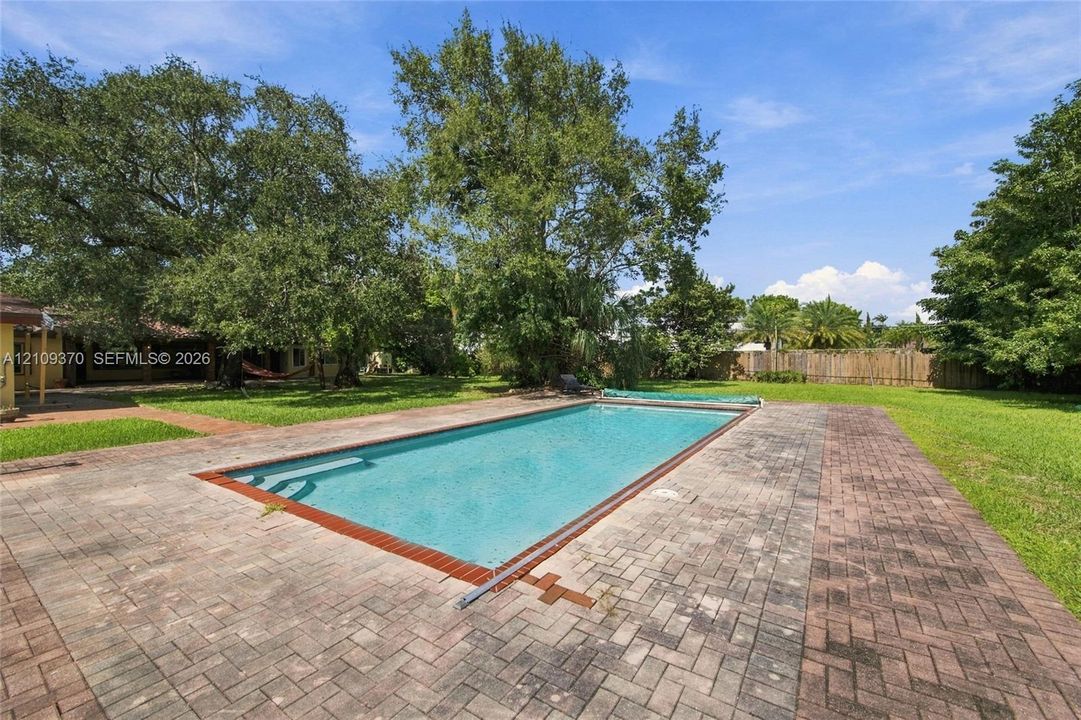 Rectangular inground pool with a brick surround, paved patio, expansive lawn, mature trees, and wooden perimeter fencing