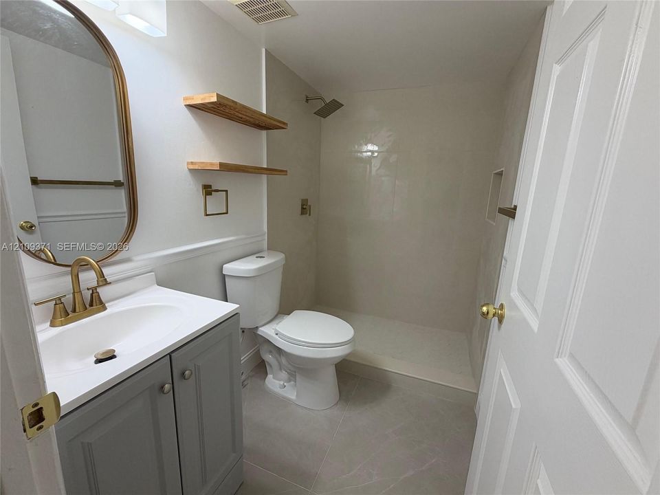 Bathroom featuring a grey vanity with a white countertop, brass-finish faucet and hardware, walk-in shower with large-format tile surround, and floating wood shelving