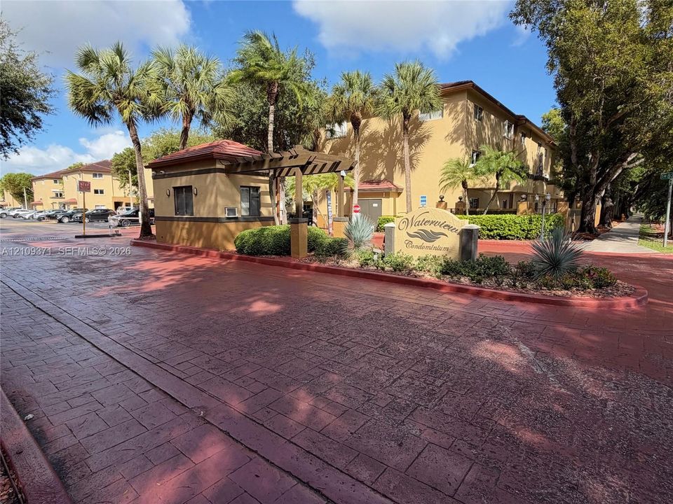 Gated entrance featuring a stamped concrete drive, an attended guard house, and mature palm landscaping