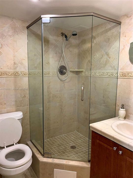 Corner glass-enclosed shower with a handheld fixture, decorative tile backsplash, integrated shelving, and neutral-tone stone-finish wall tile